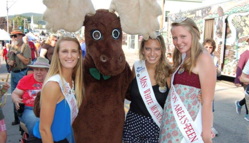 Three young women in pageant sashes pose with a person in a moose costume at a festive outdoor event.