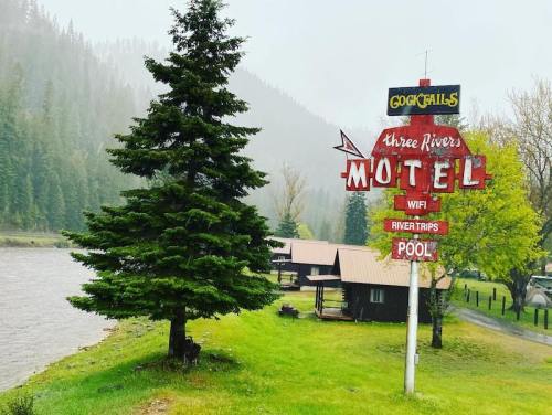 Sign for "Three Rivers Motel" by a river, surrounded by trees and misty mountains, with cabins and a pool nearby.