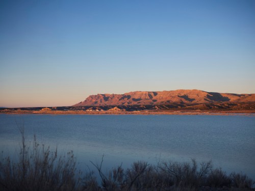A serene lake reflects the soft light of dawn, with mountains in the background under a clear blue sky.