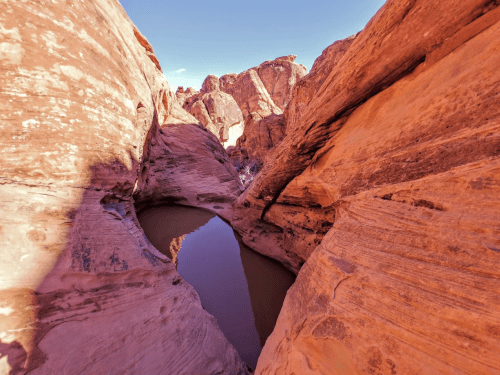 A narrow canyon with reddish rock formations and a small pool of water at the bottom, under a clear blue sky.