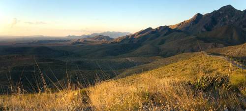 A panoramic view of rolling hills and mountains under a clear sky, with golden grass in the foreground.