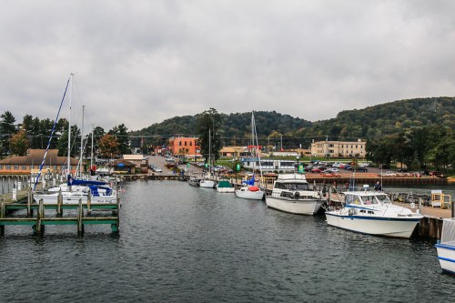 A scenic harbor with boats docked, surrounded by trees and buildings under a cloudy sky.