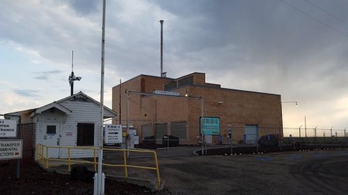 A large industrial building with a smokestack, surrounded by a fence and security gate, under a cloudy sky.