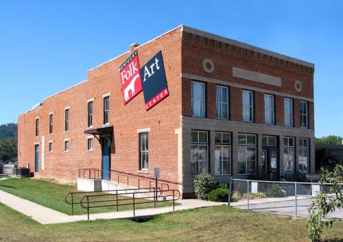Exterior view of the Folk Art Center, a brick building with large windows and a prominent sign. Green grass in foreground.