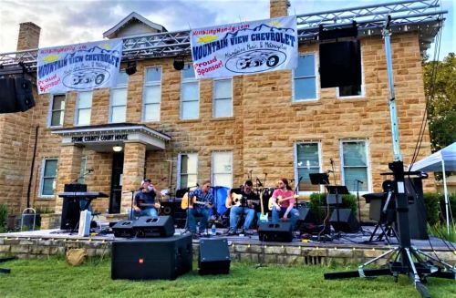 A band performs on stage in front of a stone building, with banners and sound equipment set up for an outdoor event.