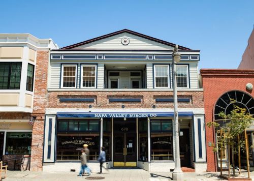 Exterior of Napa Valley Burger Co., a two-story building with large windows and a blue awning, on a sunny day.