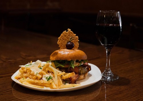 A gourmet burger topped with lettuce and a cherry, served with fries and a glass of red wine on a wooden table.