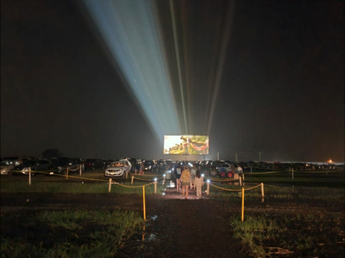 A drive-in movie scene at night, with cars parked and a large screen displaying a film, illuminated by bright beams.