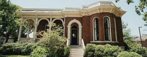 A historic brick house with arched windows and a decorative roof, surrounded by lush greenery.