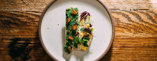 A beautifully plated dish featuring a green herb sauce, garnished with microgreens and colorful toppings on a wooden table.