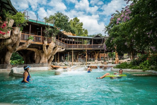 A serene pool scene with people relaxing in water, surrounded by lush greenery and a wooden structure in the background.