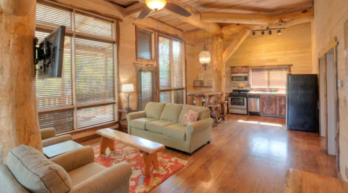 Cozy wooden cabin interior with a living area, kitchen, and large windows, featuring a sofa and rustic decor.
