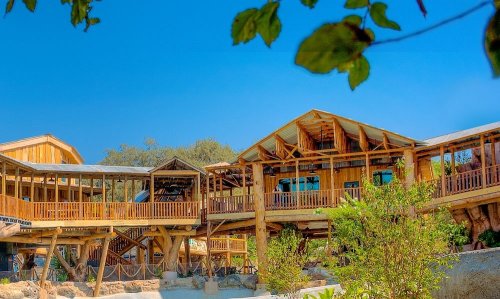 A wooden building with multiple levels, surrounded by greenery and blue skies, showcasing a rustic architectural style.