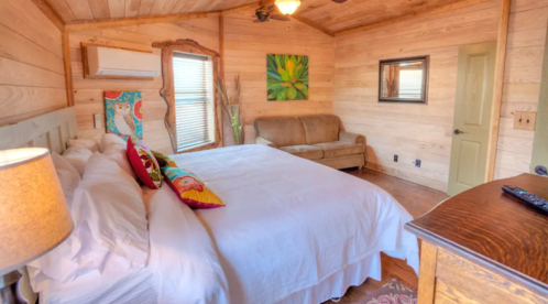 Cozy bedroom with a large bed, colorful pillows, a sofa, and wooden walls, featuring a mirror and air conditioning unit.
