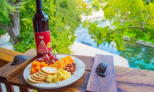 A plate of cheese, crackers, and fruit beside a bottle of wine, with a scenic view of trees and water in the background.