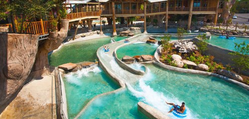 A scenic water park with winding lazy rivers, rocky landscapes, and wooden structures, featuring people floating on tubes.