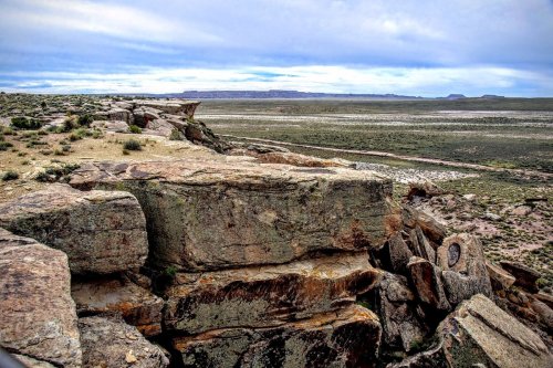 A rocky outcrop overlooks a vast, arid landscape with sparse vegetation and distant mesas under a cloudy sky.