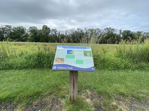 A sign about open prairies and grasslands stands in a grassy area with trees in the background under a cloudy sky.