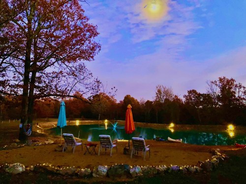 A serene evening scene by a pond, featuring lounge chairs, colorful umbrellas, and autumn foliage under a glowing moon.