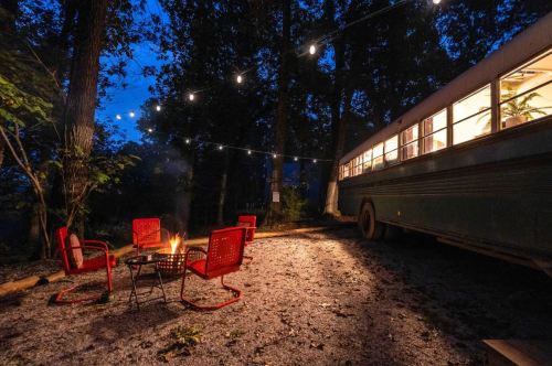 A cozy campsite at dusk with a fire pit, red chairs, and a vintage bus surrounded by trees and string lights.