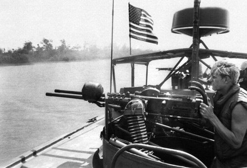 A soldier operates a machine gun on a boat, with an American flag waving in the background near a river.