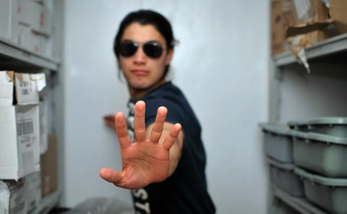A person wearing sunglasses extends their hand in a stopping gesture, standing in a narrow storage space.