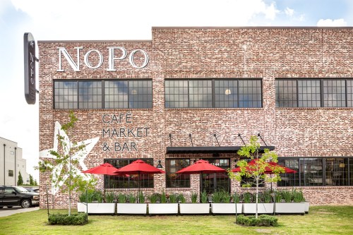 Exterior of NoPo Cafe Market & Bar, featuring a brick building, outdoor seating with red umbrellas, and greenery.