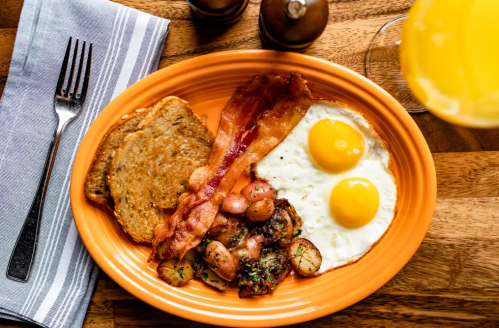 A vibrant orange plate with two sunny-side-up eggs, crispy bacon, toast, and sautéed vegetables on a wooden table.