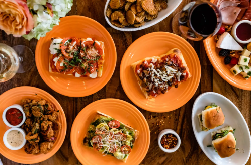 A vibrant spread of various dishes on orange plates, including pizzas, appetizers, and a glass of red wine.