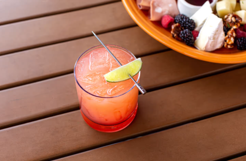 A vibrant cocktail with ice and a lime wedge, next to a platter of fruits and snacks on a wooden table.