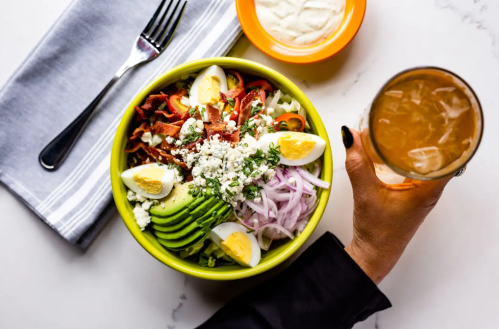A colorful salad with eggs, bacon, avocado, and onions, alongside a drink and a small bowl of sauce.