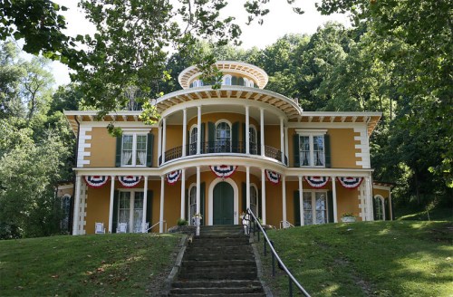 A large, historic yellow house with white trim, surrounded by trees and decorated with red, white, and blue bunting.