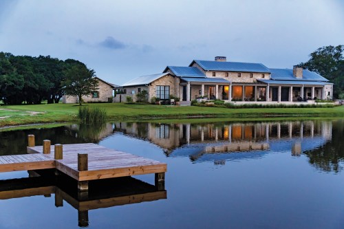 A serene lakeside view of a large stone house with a blue roof, reflecting in calm water at dusk.
