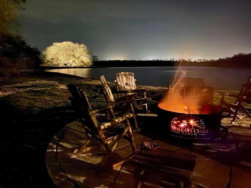 A serene lakeside scene at night with a glowing campfire and wooden chairs arranged around it.
