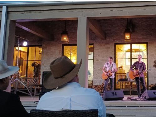 Two musicians perform on a porch, while an audience watches from the foreground, with warm lighting and a rustic setting.