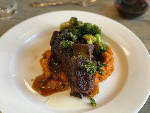 A plate of tender beef short ribs on a bed of sweet potato puree, topped with broccoli and a savory sauce.