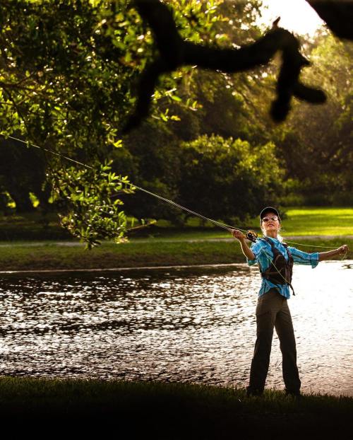 A person stands by a river, casting a fishing line with a focused expression, surrounded by lush greenery.