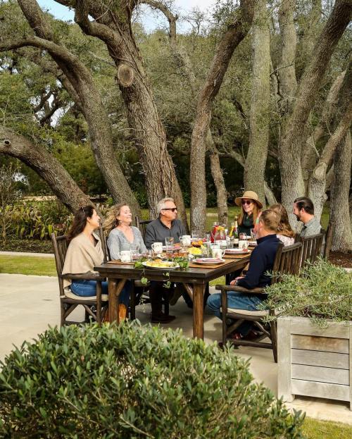 A group of people enjoying a meal outdoors at a large wooden table surrounded by trees.