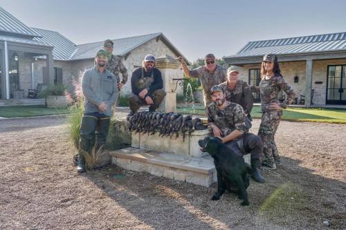 A group of hunters poses by a fountain with their hunting gear and a black dog, showcasing their recent catch of ducks.
