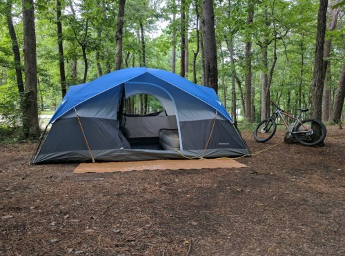 A blue and gray tent set up in a wooded area, with a bicycle parked nearby on a brown mat.