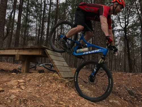 A mountain biker jumps off a wooden ramp in a forested area, showcasing action and skill on a trail.