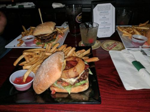 A close-up of a burger with bacon and onion rings, served with fries and dipping sauce on a wooden table.