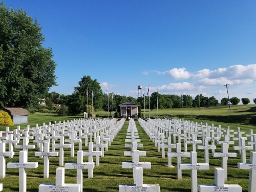 A serene cemetery with rows of white crosses under a clear blue sky, flanked by trees and flags in the background.