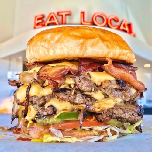 A towering burger with multiple beef patties, cheese, bacon, lettuce, and tomato, set against a bright "EAT LOCAL" sign.
