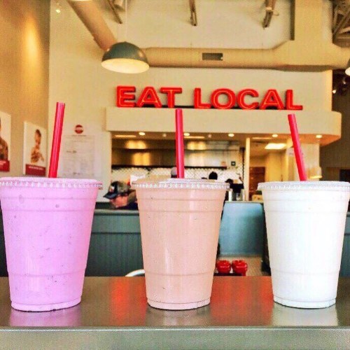Three colorful milkshakes in cups with straws, set against a backdrop featuring a sign that says "EAT LOCAL."