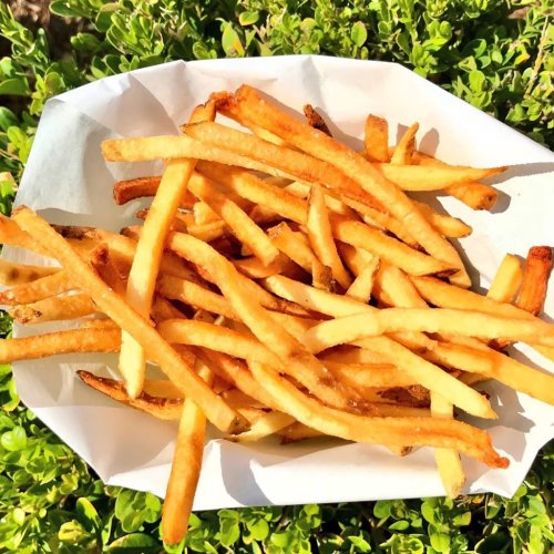 A serving of golden, crispy French fries on a white paper tray, surrounded by green foliage.