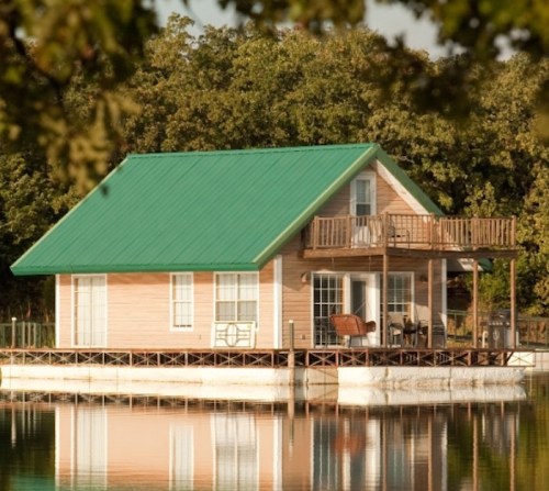 A charming lakeside house with a green metal roof and a deck, surrounded by trees and reflecting in the water.