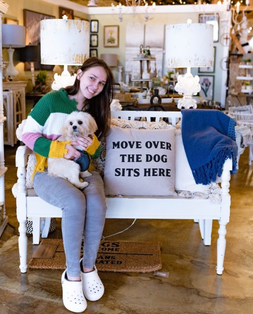 A woman sits on a bench holding a small dog, with a decorative pillow that says "MOVE OVER THE DOG SITS HERE."