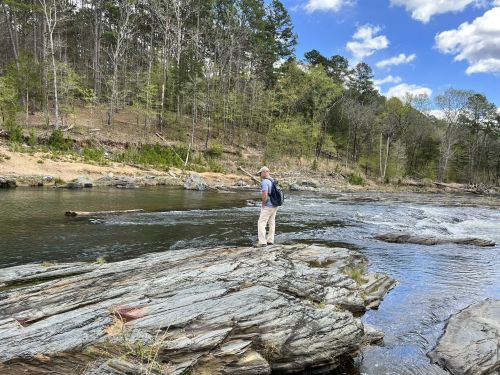 A person stands on a rocky riverbank, gazing at the water surrounded by trees and a blue sky with clouds.