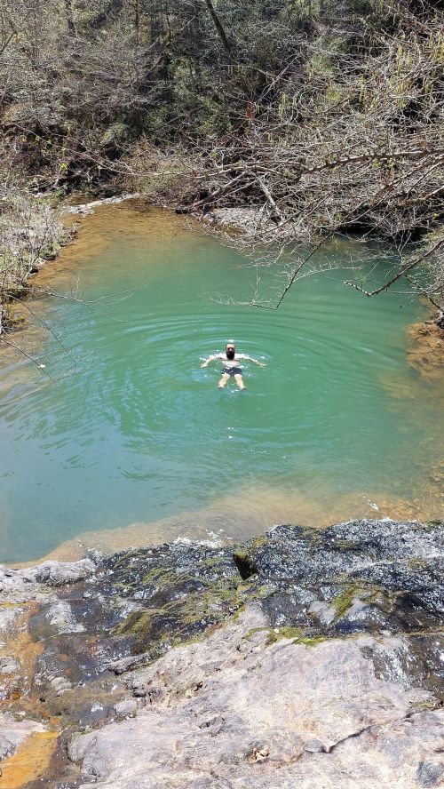 A person swims in a clear, greenish pool surrounded by rocky terrain and sparse trees.